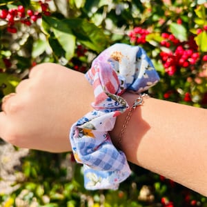 May include: A multi-patterned scrunchie with a small chain detail. The scrunchie features a mix of floral, gingham, and striped fabrics in shades of blue, white, and red. The accessory is worn on the wrist.
