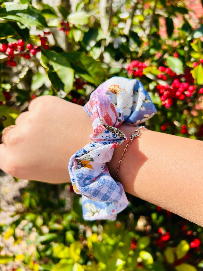 Może przedstawiać: Wzorzysta gumka do włos&oacute;w z małym łańcuszkiem. Gumka do włos&oacute;w ma mieszankę kwiatowych, w kratkę i w paski tkanin w odcieniach niebieskiego, białego i czerwonego. Akcesorium jest noszone na nadgarstku.