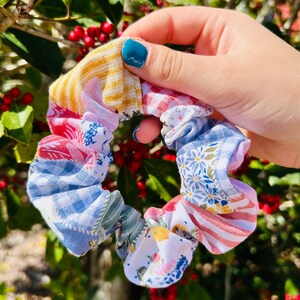 Może przedstawiać: Kolorowa gumka do włos&oacute;w w stylu patchwork z r&oacute;żnymi wzorami tkanin, w tym kratą, kwiatami i paskami. Gumka jest trzymana na tle zielonych liści i czerwonych jag&oacute;d, prezentując swoje żywe kolory i tekstury.