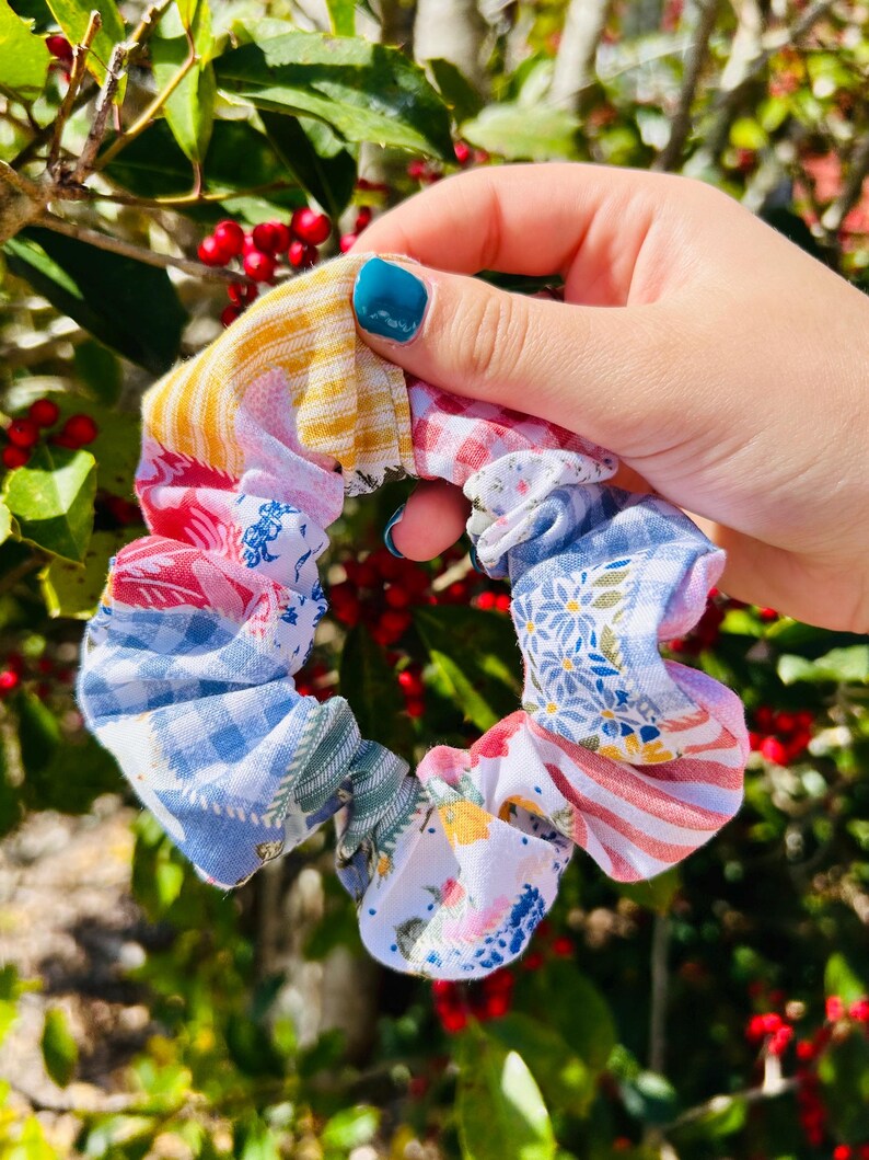 Może przedstawiać: Kolorowa gumka do włos&oacute;w w stylu patchwork z r&oacute;żnymi wzorami tkanin, w tym kratą, kwiatami i paskami. Gumka jest trzymana na tle zielonych liści i czerwonych jag&oacute;d, prezentując swoje żywe kolory i tekstury.