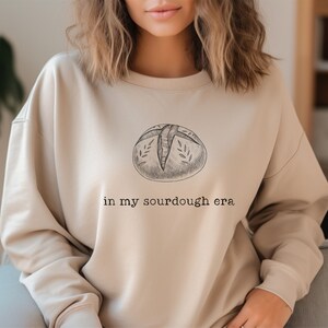 Może przedstawiać: Beżowa bluza z czarno-białym rysunkiem chleba na zakwasie i tekstem "in my sourdough era".