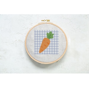 May include: A cross-stitch embroidery hoop featuring a carrot design. The carrot is orange with green leaves, set against a blue and white gingham background. The wooden hoop is round and secured with a metal clasp. The craft project is displayed on a white textured surface.