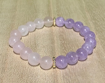 Puede incluir: Un brazalete de piedras preciosas rosa y púrpura con detalles dorados. El brazalete está hecho de cuentas redondas y está enhebrado en un cordón elástico.