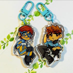 May include: Two acrylic keychains featuring cartoon characters with light blue plastic lobster clasps. The left character has red hair, blue goggles, and a yellow and blue suit. The right character has brown hair, a blue shirt, and a small black dog.