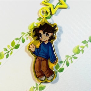 May include: A yellow acrylic keychain featuring a cartoon character with dark brown hair, wearing a blue long-sleeved sweater and brown corduroy-style pants. The character is waving one hand. The keychain has a yellow star-shaped plastic clasp.
