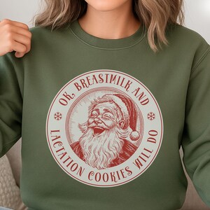 May include: A military green sweatshirt with a circular graphic featuring Santa Claus and the text "OK, Breastmilk and Lactation Cookies Will Do". The design is in red and white, with snowflake accents. The sweatshirt is a casual, long-sleeved garment.