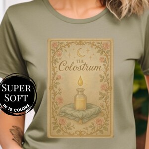 May include: Olive green t-shirt featuring a tarot card design with the text "The Colostrum". The card depicts a bottle with a drop above it, surrounded by floral elements. A circular badge reads "Super Soft in 16 Colors".