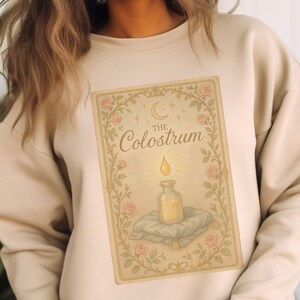 May include: Sand-colored sweatshirt featuring a vintage-style graphic. The graphic includes the text "The Colostrum" and an illustration of a bottle with a drop above it, surrounded by floral elements. The sweatshirt is a neutral color.
