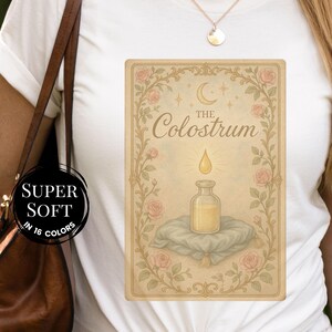 May include: White t-shirt featuring a vintage-style illustration of a bottle with a drop of liquid, the text "The Colostrum", and a floral border. The shirt also has the text "Super Soft in 16 Colors".