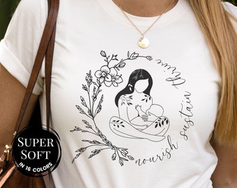 May include: White t-shirt with a black illustration of a woman breastfeeding a baby. The illustration is surrounded by floral designs and the text "nourish sustain thrive".