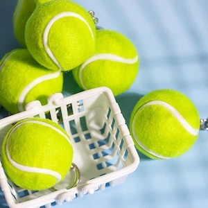 May include: A collection of bright yellow tennis ball keychains with white stripes. One keychain is inside a small white basket. The keychains are on a light blue surface.