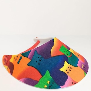May include: A colorful visor with a pattern of cats in various shades of blue, green, orange, pink, and purple. The visor is made of a lightweight fabric and has a red elastic band.