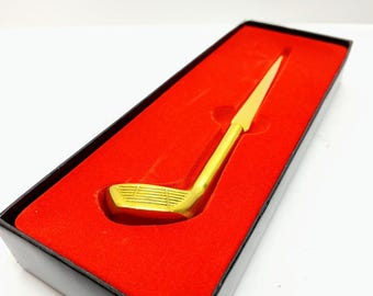 May include: A gold-colored golf club-shaped item, presented in a black box with a red velvet interior. The golf club is a detailed miniature, likely a decorative piece or a gift item.