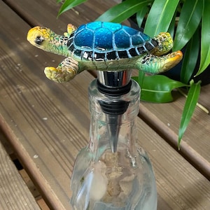 May include: A decorative bottle stopper featuring a turtle design. The turtle has a blue shell, green body, and yellow accents. The stopper is made of metal and fits into a clear glass bottle. The bottle is partially filled with a clear liquid.
