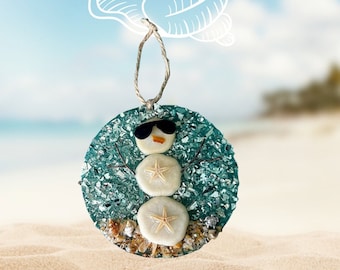 May include: A round, blue and green glass ornament with a snowman design made from sand dollars and seashells. The snowman is wearing sunglasses and has a small orange nose.