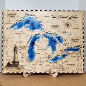May include: A wooden map of the Great Lakes, featuring detailed laser-cut topography. The map highlights the lakes with varying shades of blue, and includes a lighthouse illustration. The text "The Great Lakes" is displayed in elegant script.