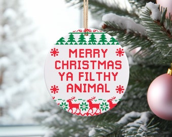 Op de afbeelding: Een witte, ronde kerstversiering met de tekst "MERRY CHRISTMAS YA FILTHY ANIMAL" in rode pixelletters. De versiering heeft een groen-witte rand met kerstbomen en rendieren. Een gouden touwtje is bevestigd om op te hangen.