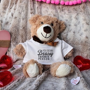 May include: A brown teddy bear wearing a white t-shirt with the text "You're my Beary Favorite Person" in black. The bear is sitting on a fluffy white blanket with red hearts and a pink heart-shaped decoration.