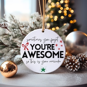Può includere: Ornamento natalizio rotondo in ceramica bianca con cordino dorato. L'ornamento presenta la scritta "sometimes you forget YOU'RE AWESOME so this is your reminder" in nero e rosso. Un fiocco rosso e una stella verde decorano l'ornamento.