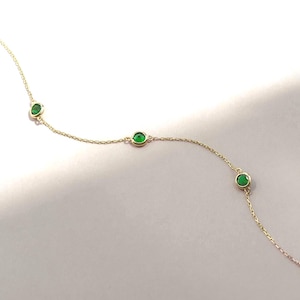 May include: A delicate gold chain necklace featuring three evenly spaced, round, vibrant green gemstones set in gold bezels. The chain is thin and metallic, with the stones positioned along the length of the strand in a simple, linear arrangement.