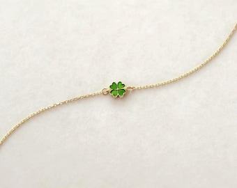 May include: A delicate gold chain bracelet featuring a small, centered four-leaf clover charm. The clover charm is filled with vibrant green enamel, creating a bright contrast against the thin, polished gold-tone metal links of the chain.