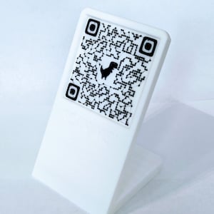 Peut inclure: Support en plastique blanc avec un code QR noir et blanc représentant un petit dinosaure noir.