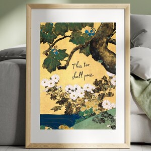May include: Framed art print featuring a Japanese-inspired illustration. The artwork displays a tree branch with green leaves and blue flowers, white chrysanthemums, and the text "This too shall pass." The background is gold.