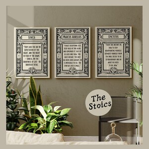 May include: Three black and white framed prints with inspirational quotes from Seneca, Marcus Aurelius, and Epictetus. The quotes are about living in the present moment and not dwelling on the past or future. The prints are arranged in a row on a wall with a light brown background. The text below the prints reads "The Stoics".