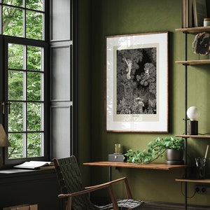 K&ouml;nnte beinhalten: Eine Schwarzwei&szlig;illustration einer Waldszene mit B&auml;umen und einem Bach. Die Illustration ist in einem braunen Holzrahmen gerahmt und h&auml;ngt an einer gr&uuml;nen Wand. Ein Holztisch mit einer Topfpflanze steht vor dem Kunstwerk.