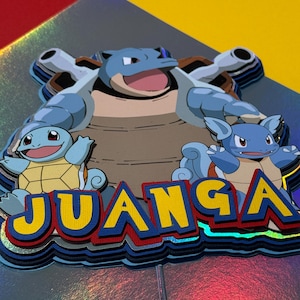 May include: A layered, colorful decoration featuring three cartoon turtle-like creatures. The top layer features a large blue turtle with cannons, and the bottom layer has the word "JUANGA" in yellow, blue, and red. The background is a mix of red, yellow, and iridescent colors.