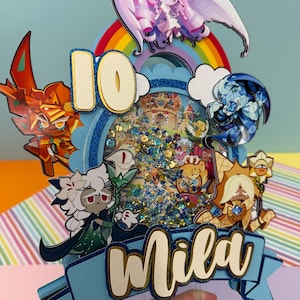 May include: A colorful cake topper with the number 10 and the name "Mila". The topper features cartoon characters, a rainbow, and a castle scene. The background is a light blue color.