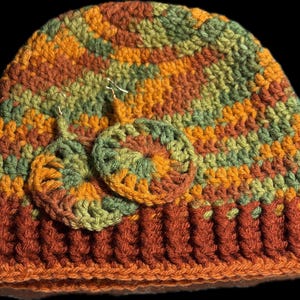 May include: A crocheted beanie hat in shades of orange, green, and brown. The hat has a striped pattern and is topped with two crocheted earrings in the same colors.