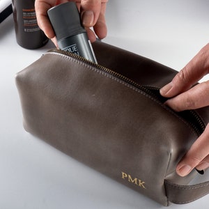 May include: A brown leather travel toiletry bag with a zipper closure and a gold zipper pull. The bag has a gold embossed monogram "PMK" on the front.