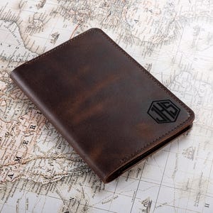 May include: A brown leather wallet with a monogram in a hexagonal shape. The wallet is placed on a vintage map. The monogram features the initials JHS. The leather has a distressed appearance.