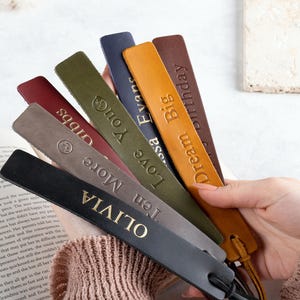 May include: A set of seven leather bookmarks with gold lettering. The bookmarks are in various colors, including brown, green, gray, and black. The text on the bookmarks includes "Olivia", "More", "Ten Love You", "Dream Big", "Lisa Evans", "One You", and "Midds".