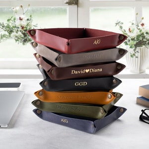 May include: A stack of eight leather trays in various colors, each with a personalized inscription in gold lettering. The trays are stacked on a white surface with a laptop, a phone, a book, and glasses.