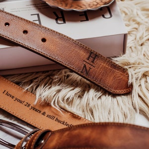 May include: Two brown leather belts with engraved text. The top belt has the initials "H/N" engraved on it. The bottom belt has the text "I love you! 28 years & still buckled" engraved on it.