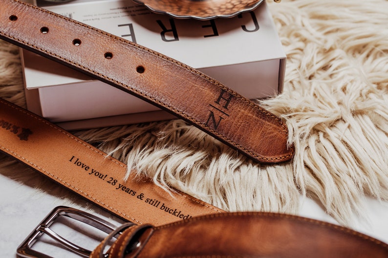 May include: Two brown leather belts with engraved text. The top belt has the initials "H/N" engraved on it. The bottom belt has the text "I love you! 28 years & still buckled" engraved on it.