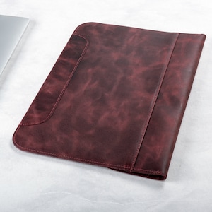 May include: A burgundy leather laptop sleeve with a flap closure and a subtle textured finish.