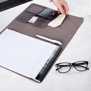 Puede incluir: Un portafolio de cuero gris con un bloc de notas, un bol&iacute;grafo y un tel&eacute;fono inteligente en su interior. El portafolio tiene un bolsillo para tarjetas y una solapa que se cierra con un broche a presi&oacute;n.
