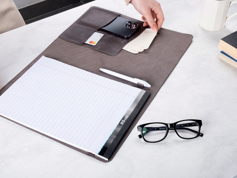Puede incluir: Un portafolio de cuero gris con un bloc de notas, un bol&iacute;grafo y un tel&eacute;fono inteligente en su interior. El portafolio tiene un bolsillo para tarjetas y una solapa que se cierra con un broche a presi&oacute;n.