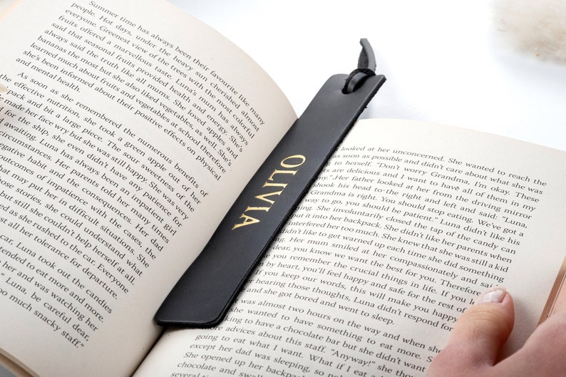 May include: A black leather bookmark with gold lettering that reads "OLIVIA". The bookmark is laying on a book with text about the benefits of eating fruits and vegetables.