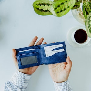 May include: A blue leather wallet, open to display credit cards. The wallet is held in two hands, with a cup of coffee and a plant in the background. The wallet has multiple card slots and a clear ID window.