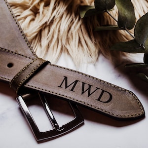 May include: A brown leather belt with a silver buckle. The belt is personalised with the initials "MWD" engraved in the leather.
