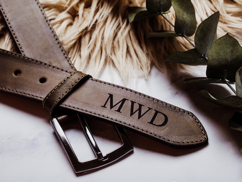 May include: A brown leather belt with a silver buckle. The belt is personalised with the initials "MWD" engraved in the leather.