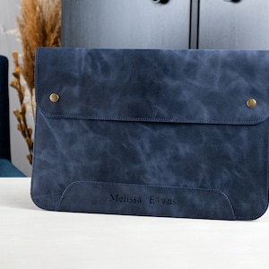 May include: A blue leather laptop sleeve with a flap closure and two brass snaps. The sleeve has a personalized inscription that reads "Melissa Evans".