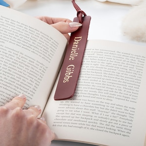May include: A burgundy leather bookmark with gold lettering that reads "Danielle Gibbs". The bookmark is being held open by a hand, revealing a page of a book.