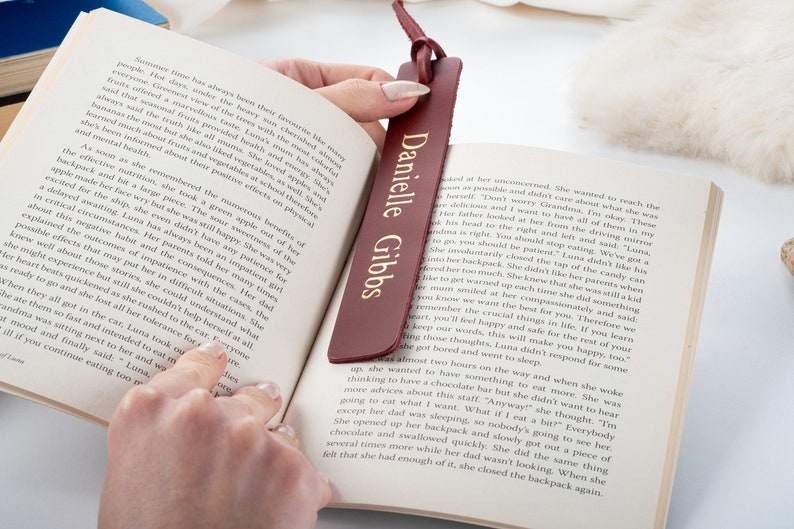 May include: A burgundy leather bookmark with gold lettering that reads "Danielle Gibbs". The bookmark is being held open by a hand, revealing a page of a book.
