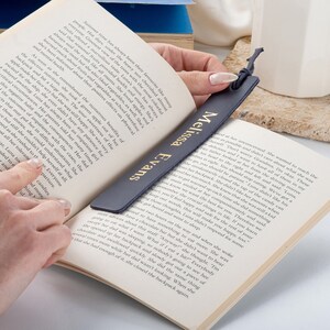 May include: A dark blue leather bookmark with gold lettering that reads "Melissa Evans". The bookmark is being held by a hand, and is placed in a book.