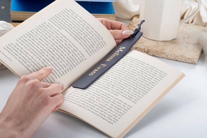 May include: A dark blue leather bookmark with gold lettering that reads "Melissa Evans". The bookmark is being held by a hand, and is placed in a book.